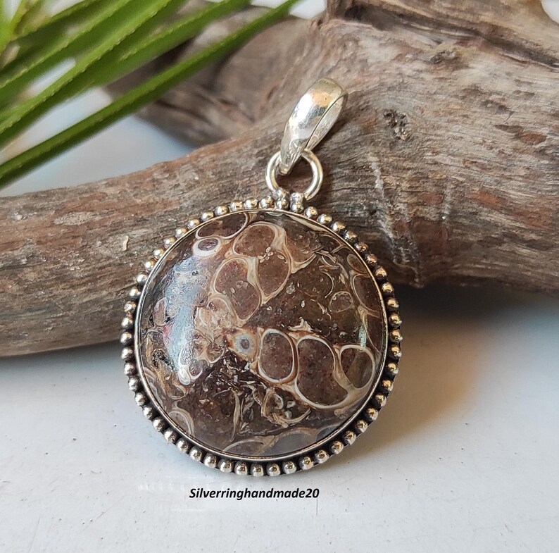 May include: A silver pendant with a round, brown and white agate stone. The stone has a marbled pattern. The pendant is on a wooden surface.