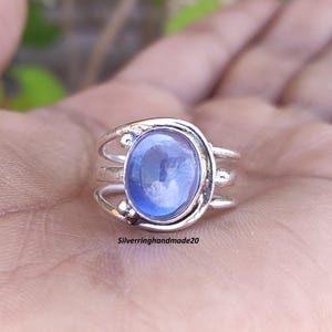 Puede incluir: Un anillo de plata con una piedra ovalada azul engastada en un diseño de triple banda. El anillo tiene un estilo simple y elegante.