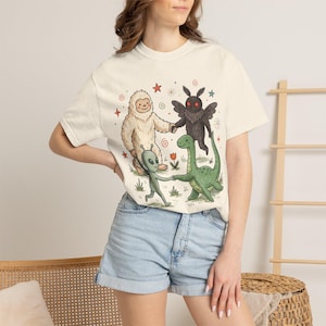 May include: A cream-colored short-sleeve t-shirt featuring a graphic illustration of a white yeti, a black moth-like creature, a green alien, and a green long-necked dinosaur holding hands in a circle, surrounded by small stars and flowers.