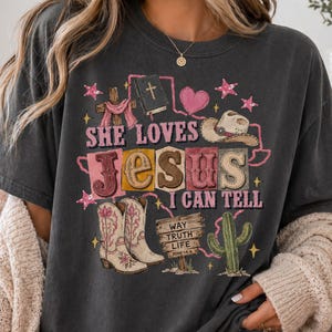 May include: A charcoal gray t-shirt featuring a graphic print with the text "SHE LOVES JESUS I CAN TELL". The design includes a cross, a Bible, a heart, a cowboy hat, a pair of cowboy boots, a cactus, and a sign reading "WAY TRUTH LIFE".