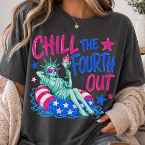 May include: A charcoal gray short-sleeve t-shirt featuring a graphic of the Statue of Liberty wearing sunglasses and relaxing in a red, white, and blue striped pool float while holding a drink. The text reads CHILL THE FOURTH OUT in pink and blue.