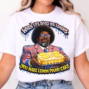 May include: White t-shirt featuring a graphic of a man in a suit with an American flag design, holding a lemon pound cake. The text reads "WHEN LIFE GIVES YOU LEMONS YOU MAKE LEMON POUND CAKE."
