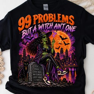 May include: A black t-shirt featuring a graphic of a green-skinned witch with curly hair, sunglasses, and a hat, sitting on a tombstone. The text reads 99 PROBLEMS BUT A WITCH AIN'T ONE and FLY HIGH STAY BAD NO CURSES JUST VIBES.