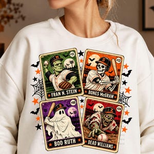 May include: A white crewneck sweatshirt featuring a graphic print of four vintage-style baseball cards. The cards depict a green monster, a skeleton, a ghost, and a zombie playing baseball, labeled Fran N. Stein, Bones McGraw, Boo Ruth, and Dead Williams.
