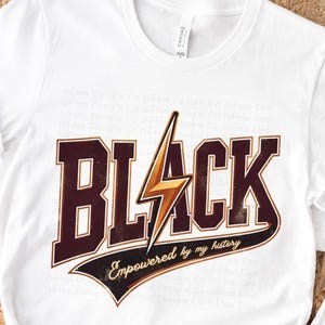 May include: White t-shirt with a graphic print of the word "BLACK" in brown letters with a gold lightning bolt. The text "Empowered by my history" is printed in a curved line below the word "BLACK".