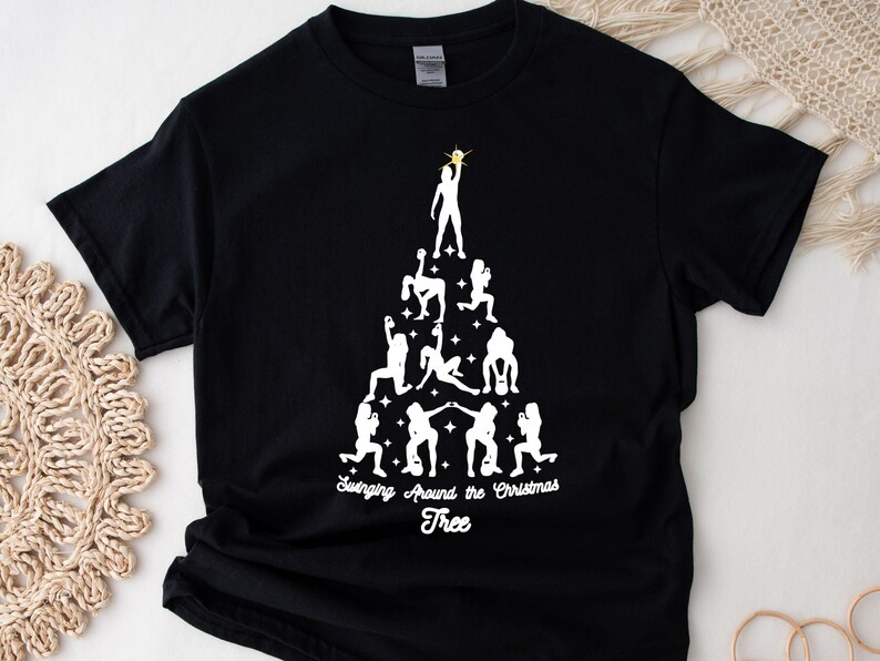 May include: Black t-shirt with a white silhouette of a Christmas tree made up of women in various exercise poses. The text "Swinging Around the Christmas Tree" is printed below the tree.