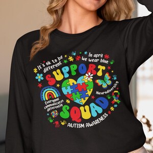May include: Black long-sleeve shirt with a colorful graphic design featuring a puzzle piece heart, rainbow, butterfly, and the text "It's ok to be different", "Support Squad", "In April we wear blue", "Everyone communicates differently", "Neurodiversity", and "Autism Awareness".