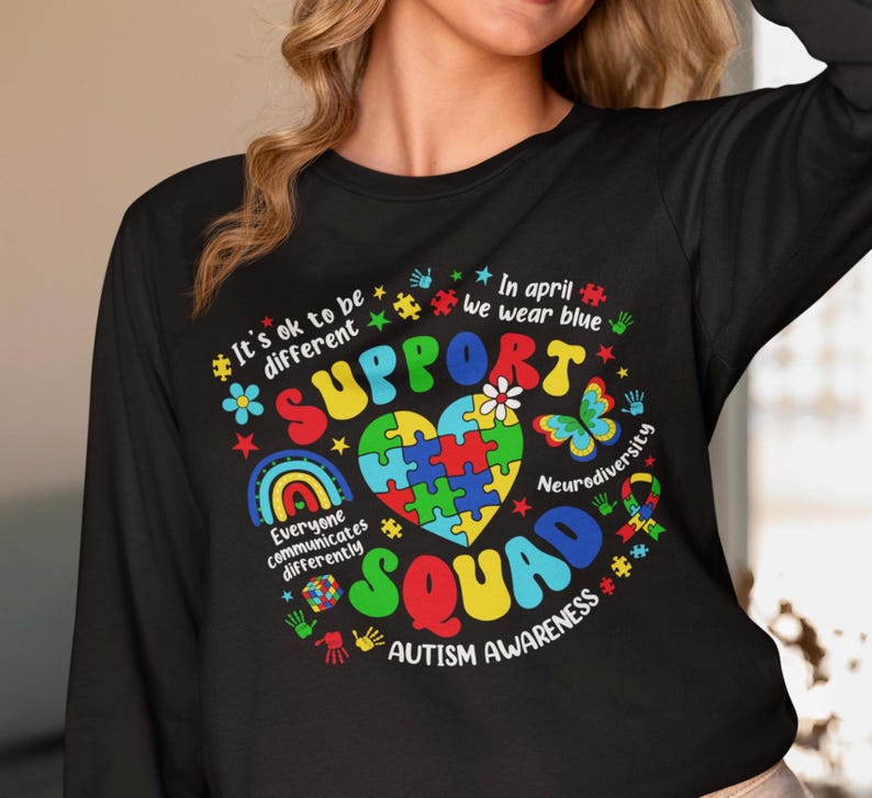May include: Black long-sleeve shirt with a colorful graphic design featuring a puzzle piece heart, rainbow, butterfly, and the text "It's ok to be different", "Support Squad", "In April we wear blue", "Everyone communicates differently", "Neurodiversity", and "Autism Awareness".