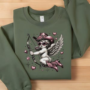 May include: Green sweatshirt with a graphic print of a pink and white cherub wearing a cowboy hat and sunglasses, holding a bow and arrow. The cherub is surrounded by pink hearts and stars.