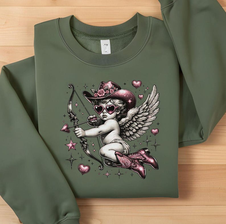 May include: Green sweatshirt with a graphic print of a pink and white cherub wearing a cowboy hat and sunglasses, holding a bow and arrow. The cherub is surrounded by pink hearts and stars.