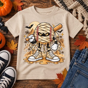 May include: A beige short-sleeve t-shirt featuring a graphic of a baseball wrapped in mummy bandages, holding a wooden baseball bat. The character wears white sneakers and stands in a desert landscape with palm trees, pyramids, and a full moon.