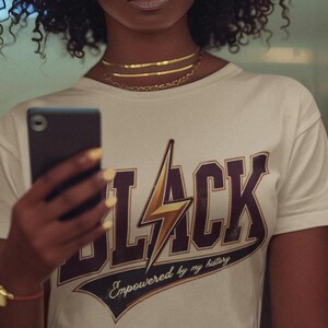 May include: A woman wearing a white t-shirt with a black and gold graphic that says "BLACK" with a lightning bolt and the text "Empowered by my history".