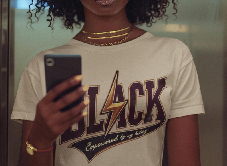 May include: A woman wearing a white t-shirt with a black and gold graphic that says "BLACK" with a lightning bolt and the text "Empowered by my history".