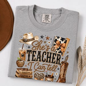 May include: A grey t-shirt featuring a graphic print with a cowboy hat, a leopard print apple, a stack of books labeled Teach and Inspire, a coffee cup, a notebook, and a cowboy boot. The text reads She's a TEACHER I Can tell.