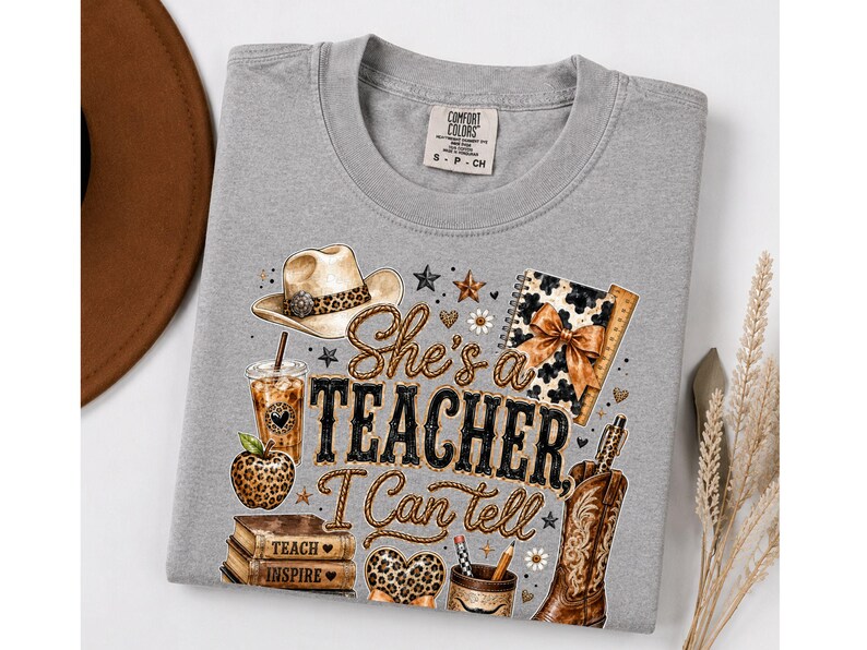 May include: A grey t-shirt featuring a graphic print with a cowboy hat, a leopard print apple, a stack of books labeled Teach and Inspire, a coffee cup, a notebook, and a cowboy boot. The text reads She's a TEACHER I Can tell.