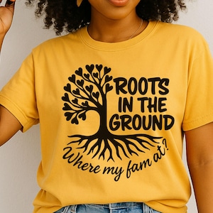 May include: Mustard yellow t-shirt with a black tree graphic. The tree has heart-shaped leaves and the text "ROOTS IN THE GROUND Where my fam at?" in a stylized font. The shirt is a crew neck.