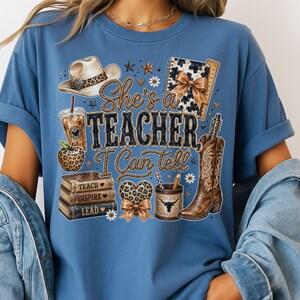 May include: A blue t-shirt featuring a graphic print with a cowboy hat, a leopard print apple, a stack of books labeled Teach, Inspire, Lead, a heart, a pencil holder, and a cowboy boot. The text reads She's a TEACHER, I Can tell.