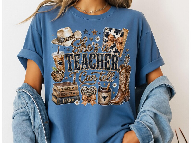 May include: A blue t-shirt featuring a graphic print with a cowboy hat, a leopard print apple, a stack of books labeled Teach, Inspire, Lead, a heart, a pencil holder, and a cowboy boot. The text reads She's a TEACHER, I Can tell.