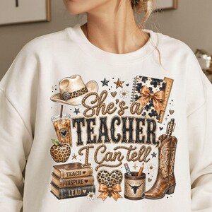 May include: A cream-colored sweatshirt featuring a graphic print with the text "She's a TEACHER, I Can tell". The design includes a cowboy hat, a leopard print apple, a stack of books labeled "TEACH, INSPIRE, LEAD", a coffee cup, and a cowboy boot.