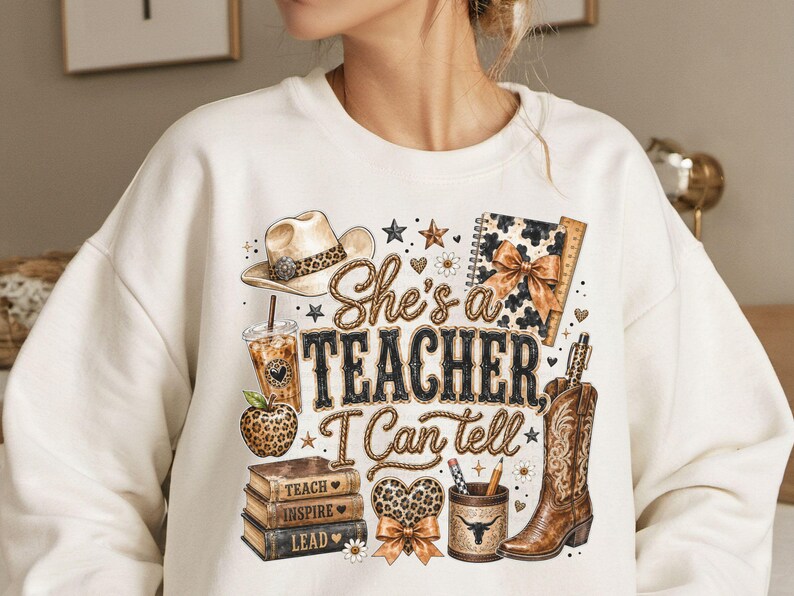 May include: A cream-colored sweatshirt featuring a graphic print with the text "She's a TEACHER, I Can tell". The design includes a cowboy hat, a leopard print apple, a stack of books labeled "TEACH, INSPIRE, LEAD", a coffee cup, and a cowboy boot.