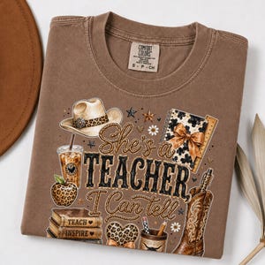 May include: A brown t-shirt featuring a graphic print with the text "She's a TEACHER I Can tell". The design includes a cowboy hat, a leopard print apple, a stack of books labeled "TEACH INSPIRE", a coffee cup, a notebook, and a cowboy boot.