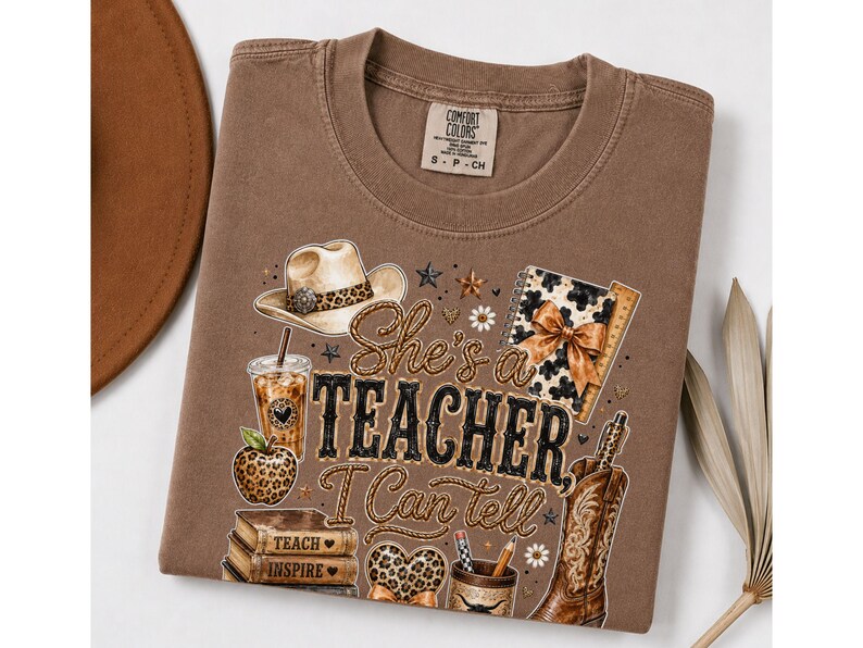 May include: A brown t-shirt featuring a graphic print with the text "She's a TEACHER I Can tell". The design includes a cowboy hat, a leopard print apple, a stack of books labeled "TEACH INSPIRE", a coffee cup, a notebook, and a cowboy boot.