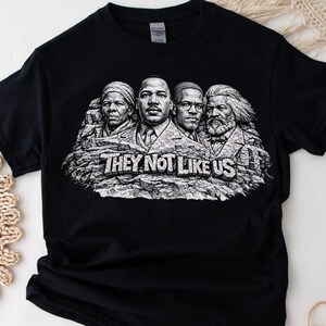 以下が含まれることがあります： 黒いTシャツ。白の「THEY NOT LIKE US」の文字と、岩の彫刻に彫られた4人の歴史上の人物のグレースケールグラフィックが特徴です。デザインはラシュモア山に似ています。シャツは柔らかい素材で作られています。