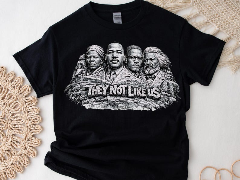 以下が含まれることがあります： 黒いTシャツ。白の「THEY NOT LIKE US」の文字と、岩の彫刻に彫られた4人の歴史上の人物のグレースケールグラフィックが特徴です。デザインはラシュモア山に似ています。シャツは柔らかい素材で作られています。