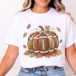 May include: A white short-sleeve t-shirt featuring a central graphic of a pumpkin designed with the texture and white laces of an American football. The pumpkin is surrounded by scattered brown and orange autumn leaves and a small white bow.
