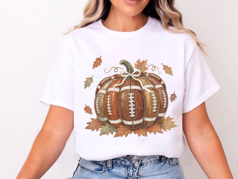 May include: A white short-sleeve t-shirt featuring a central graphic of a pumpkin designed with the texture and white laces of an American football. The pumpkin is surrounded by scattered brown and orange autumn leaves and a small white bow.