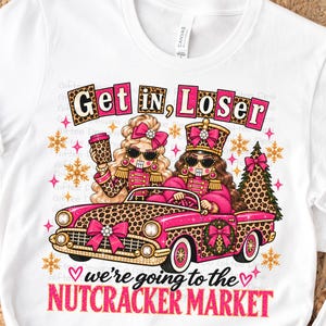 May include: A white t-shirt featuring a graphic of two nutcracker characters in a pink leopard-print convertible car. The text reads Get in, Loser we're going to the NUTCRACKER MARKET. The design includes gold snowflakes, a Christmas tree, and pink bows.