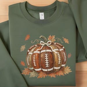 May include: An olive green crewneck sweatshirt featuring a central graphic of a pumpkin designed with the texture and white laces of a football. The pumpkin is tied with a white string bow and surrounded by scattered orange and brown autumn leaves.