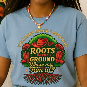 May include: A light blue t-shirt with a colorful graphic that reads "WILLIAMS FAMILY REUNION ROOTS IN THE GROUND Where my Fam at?". The graphic includes a red cowboy hat, boots, and a tree design. A beaded necklace is worn.