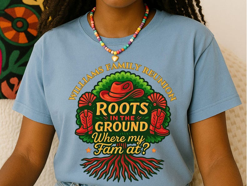 May include: A light blue t-shirt with a colorful graphic that reads "WILLIAMS FAMILY REUNION ROOTS IN THE GROUND Where my Fam at?". The graphic includes a red cowboy hat, boots, and a tree design. A beaded necklace is worn.