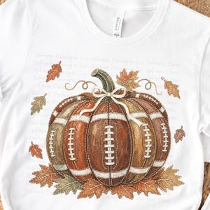 May include: A white short-sleeve t-shirt featuring a graphic of a pumpkin constructed from brown football panels with white laces. The pumpkin is decorated with a white string bow on its stem and surrounded by scattered brown and orange autumn leaves.