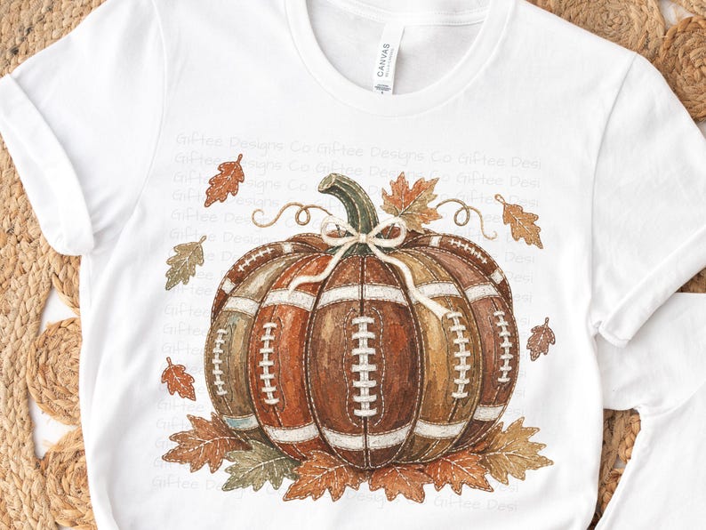 May include: A white short-sleeve t-shirt featuring a graphic of a pumpkin constructed from brown football panels with white laces. The pumpkin is decorated with a white string bow on its stem and surrounded by scattered brown and orange autumn leaves.
