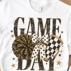 Può includere: Una t-shirt bianca a maniche corte con una stampa grafica che riporta il testo GAME DAY in lettere nere dall'aspetto usurato. Il design include un pon-pon con stampa leopardata, un fiocco color crema a pois neri, un megafono a scacchi bianco e nero e stelle dorate.