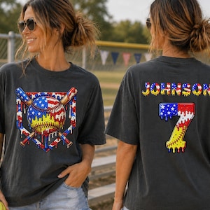 May include: A charcoal gray t-shirt featuring a front graphic of a baseball and two crossed bats over a home plate, all with a melting American flag pattern. The back displays the name JOHNSON and the number 7 in the same melting flag style.