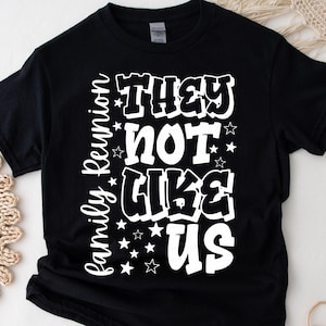 May include: Black t-shirt with the white text "THEY NOT LIKE US" in a bold, bubble font. The words "Family Reunion" are written vertically on the left side. Small white stars are scattered around the text. A casual, comfortable tee.