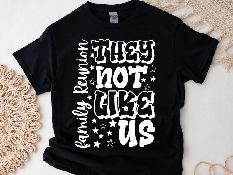 May include: Black t-shirt with the white text "THEY NOT LIKE US" in a bold, bubble font. The words "Family Reunion" are written vertically on the left side. Small white stars are scattered around the text. A casual, comfortable tee.