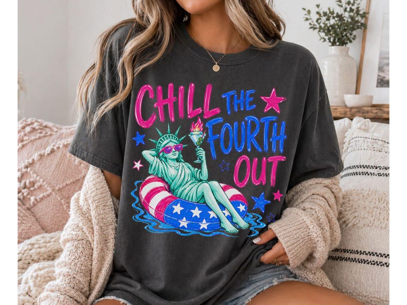 May include: A charcoal gray short-sleeve t-shirt featuring a graphic of the Statue of Liberty wearing sunglasses and relaxing in a red, white, and blue striped pool float while holding a drink. The text reads CHILL THE FOURTH OUT in pink and blue.