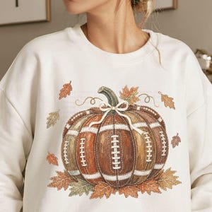May include: A cream-colored crewneck sweatshirt featuring a central graphic of a pumpkin designed with the texture and lacing of a brown leather football. The pumpkin is accented with a white bow and surrounded by scattered orange and brown autumn leaves.