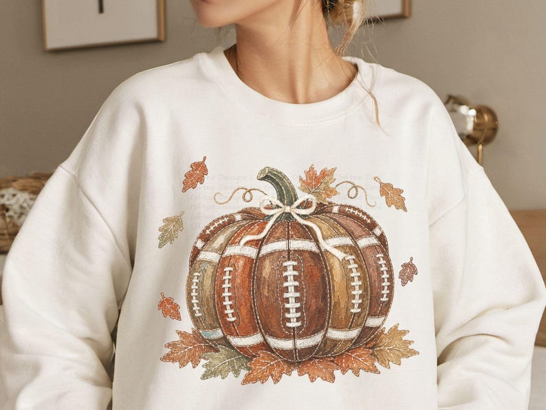 May include: A cream-colored crewneck sweatshirt featuring a central graphic of a pumpkin designed with the texture and lacing of a brown leather football. The pumpkin is accented with a white bow and surrounded by scattered orange and brown autumn leaves.