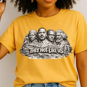 以下が含まれることがあります： マスタードイエローのTシャツ。4人の歴史上の人物が描かれたグラフィックで、マウントラッシュモアに似ており、「THEY NOT LIKE US」の文字が大胆なフォントで書かれています。柔らかい素材で作られています。