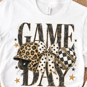 Puede incluir: Una camiseta blanca de manga corta con un estampado gráfico que incluye el texto "GAME DAY" en letras negras desgastadas. El diseño presenta un casco de sóftbol con estampado de leopardo, un lazo blanco con lunares negros y una pelota de sóftbol a cuadros.