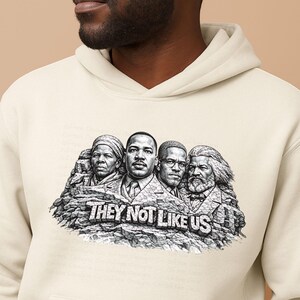 以下が含まれることがあります： クリーム色のパーカー。4人の歴史上の人物の白黒グラフィックと「THEY NOT LIKE US」の文字が特徴です。グラフィックは岩の背景に配置されています。