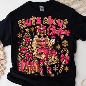 Può includere: Una t-shirt nera con una stampa grafica che riporta il testo "Nuts about Christmas". Il design include un personaggio biondo vestito da schiaccianoci, un albero di Natale decorato, pacchetti regalo, un bastoncino di zucchero e fiocchi in rosa e leopardato.