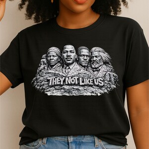以下が含まれることがあります： 歴史上の人物4人の白黒グラフィックが特徴の黒いTシャツ。記念碑の彫刻に似ています。「THEY NOT LIKE US」の文字が下にあります。柔らかい素材のシャツです。