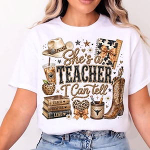 May include: A white t-shirt featuring a graphic print with the text "She's a TEACHER I Can tell". The design includes a cowboy hat, a leopard print apple, a stack of books labeled "TEACH", "INSPIRE", "LEAD", a pencil holder, and a cowboy boot.