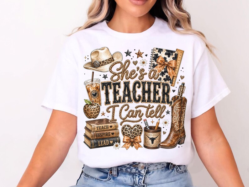 May include: A white t-shirt featuring a graphic print with the text "She's a TEACHER I Can tell". The design includes a cowboy hat, a leopard print apple, a stack of books labeled "TEACH", "INSPIRE", "LEAD", a pencil holder, and a cowboy boot.